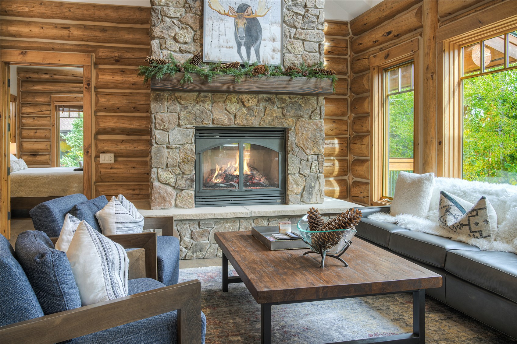 Log-cabin living room with a stone fireplace, wood walls, blue armchairs, and large windows.