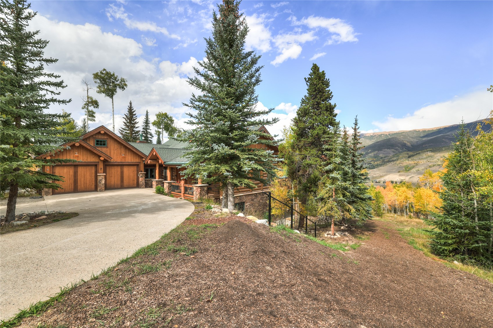 Wood-and-stone mountain home with a curved driveway, evergreen trees, and autumn mountain views.