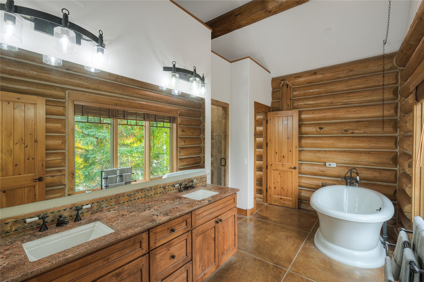 Rustic log-cabin bathroom with a long double vanity, large window, and white freestanding tub.
