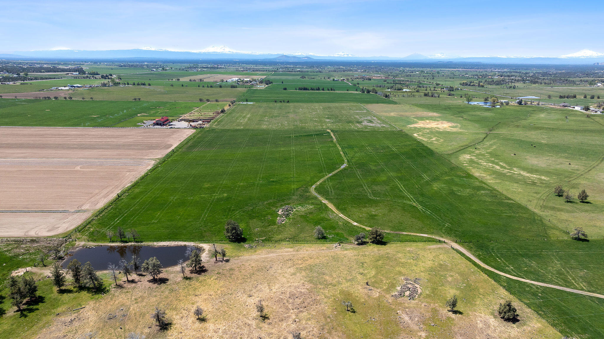 Aerial view of green ranch fields with a foreground pond and snow-capped mountains beyond.