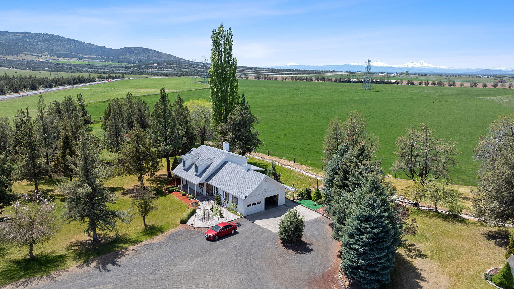 Aerial view of a white home beside a green pasture, with mature trees and snow-capped mountains beyond.