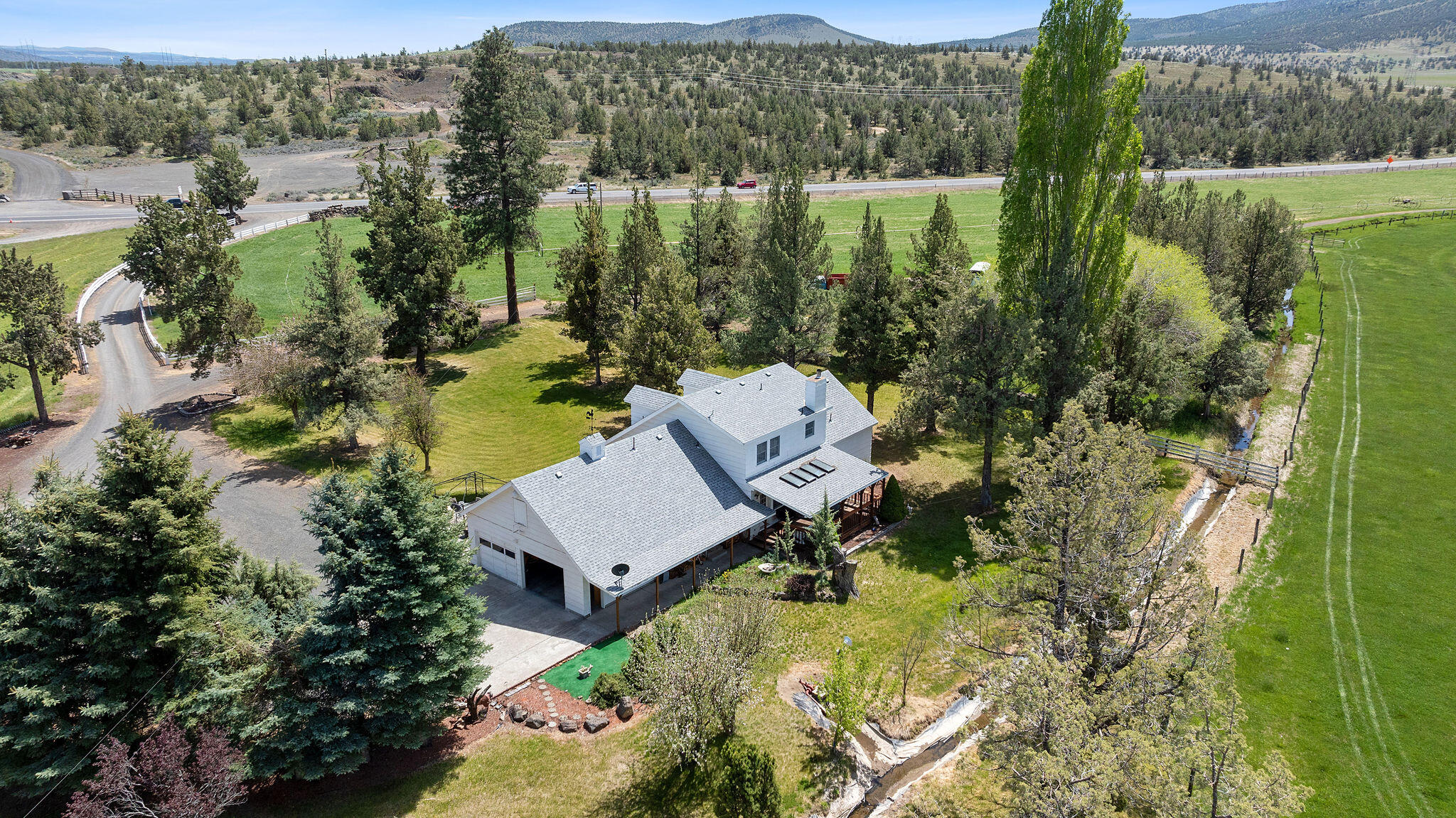 Aerial view of a white rural home among mature trees, green fields and distant hills.