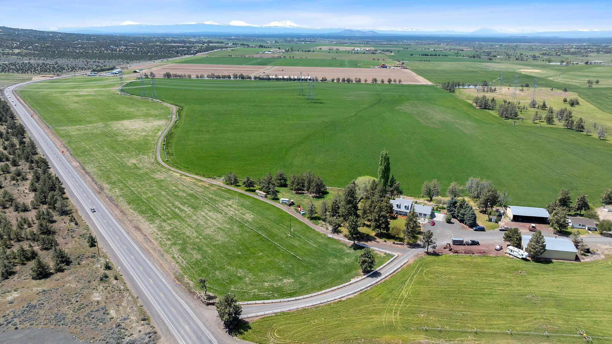 Aerial ranch view with green fields, a farmhouse, outbuildings, highway frontage, and distant snow-capped mountains.