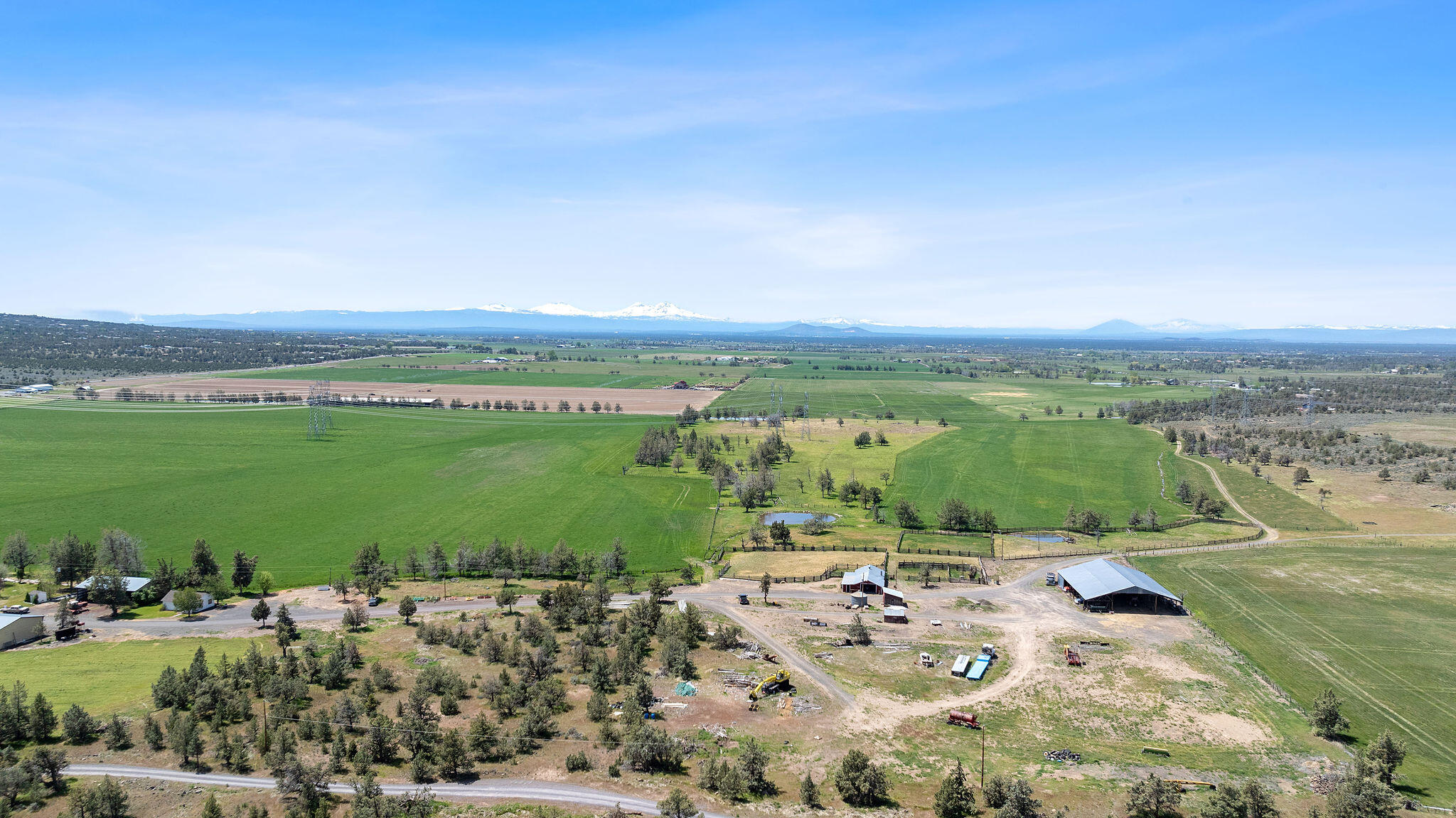 Aerial ranch view across green fields and fenced paddocks toward snow-capped mountains.