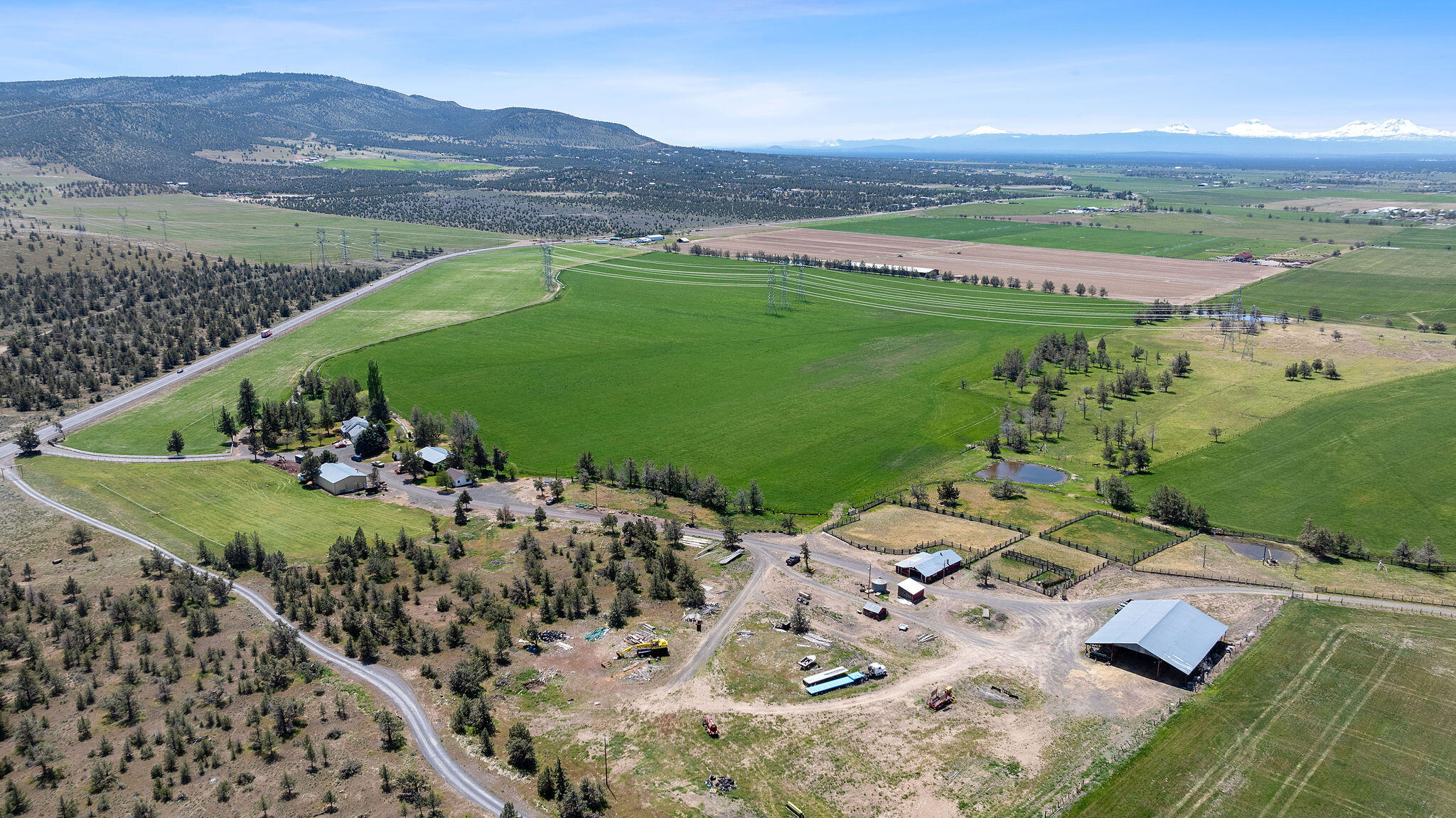 Aerial ranch view with broad green fields, fenced paddocks, a pond, and snow-capped mountains.