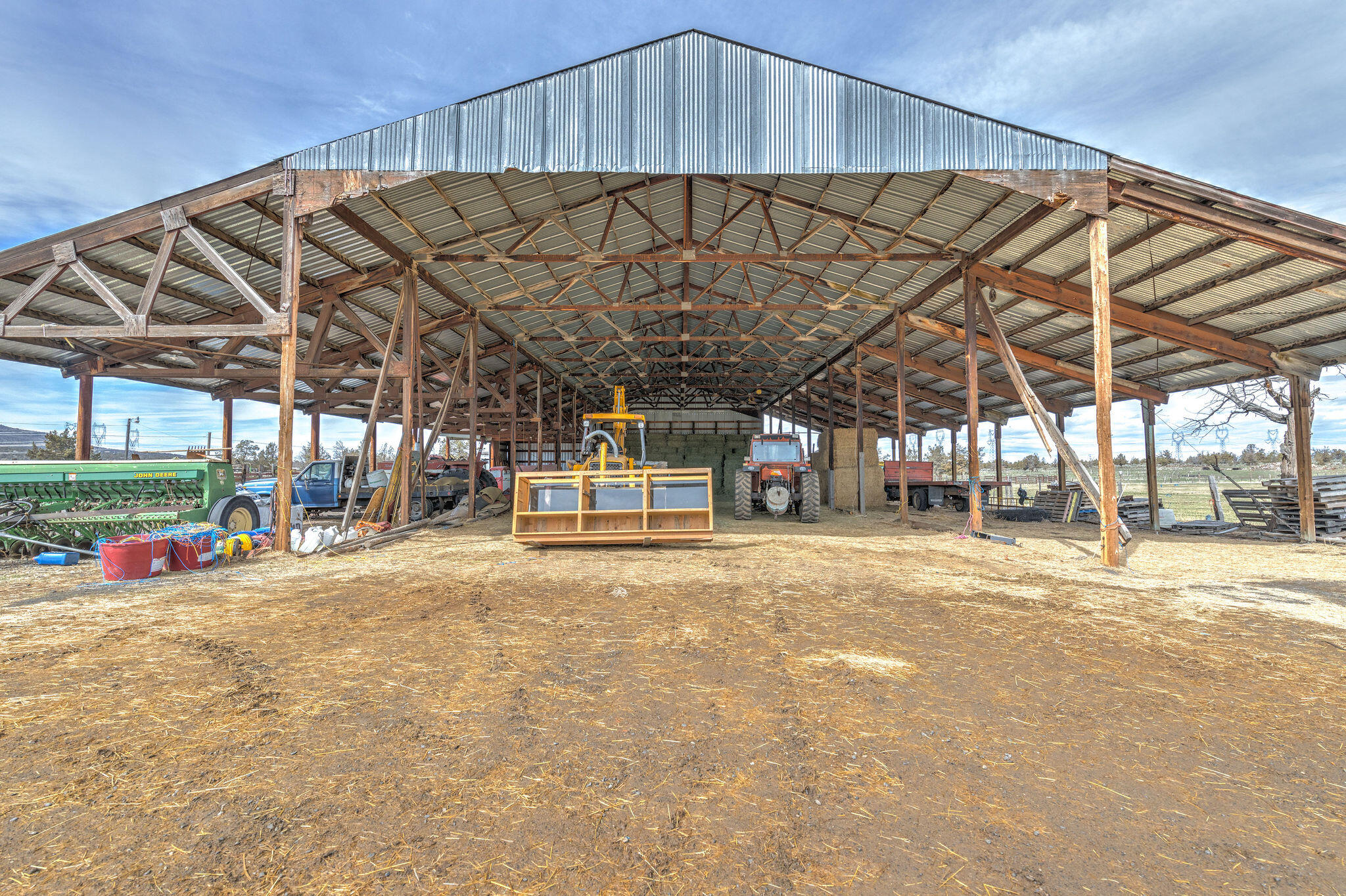 Open-sided farm shed with exposed timber trusses, tractors, and equipment on a straw-covered dirt floor.