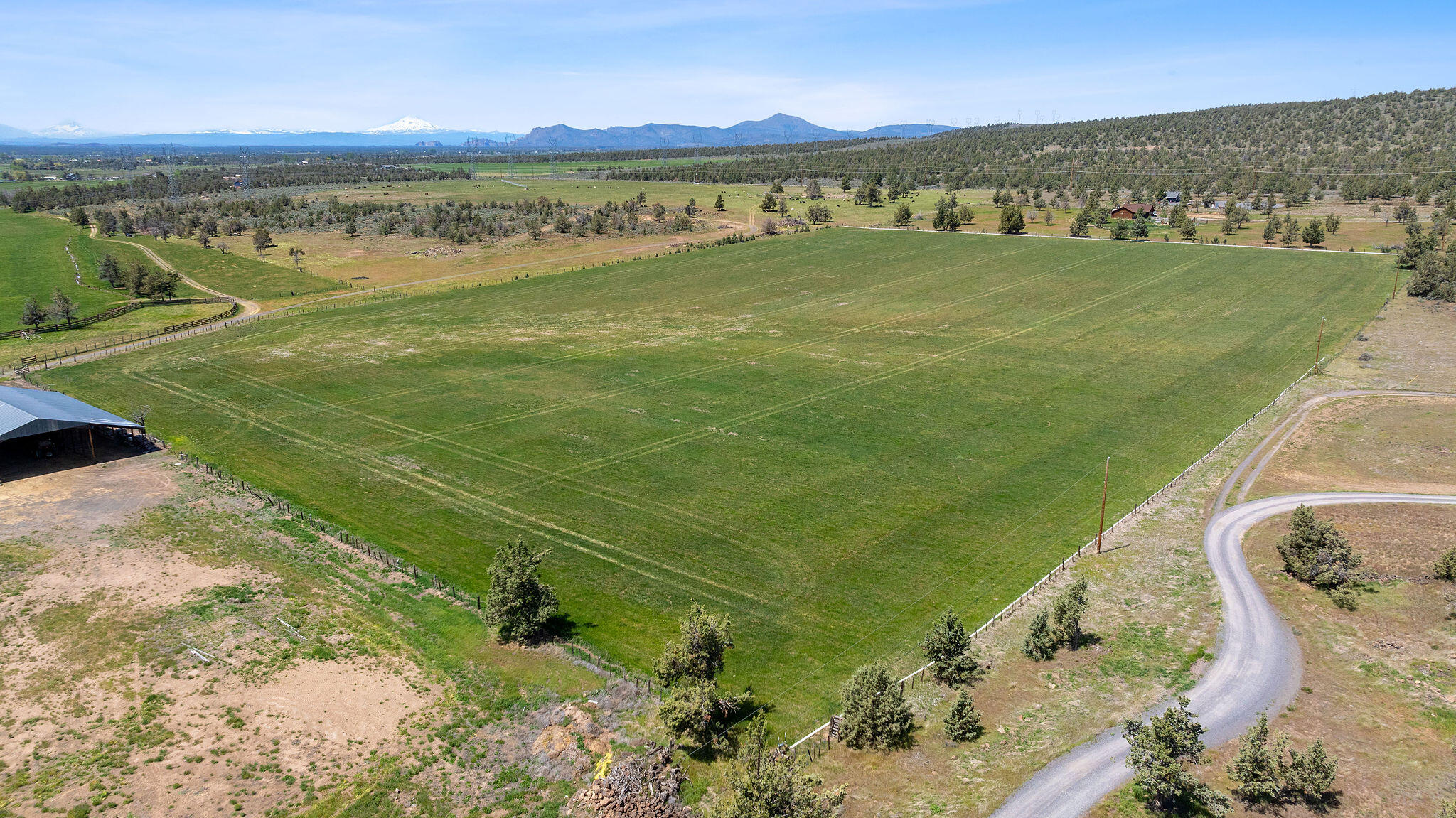 Aerial view of a fenced green field, barn, and snow-capped mountains in the distance.