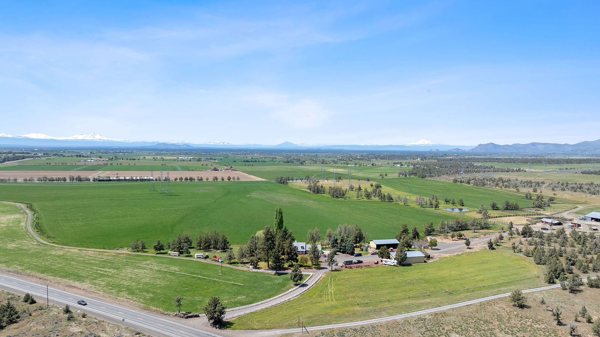 Aerial view of green fields and farm buildings, with a highway and snow-capped mountains beyond.