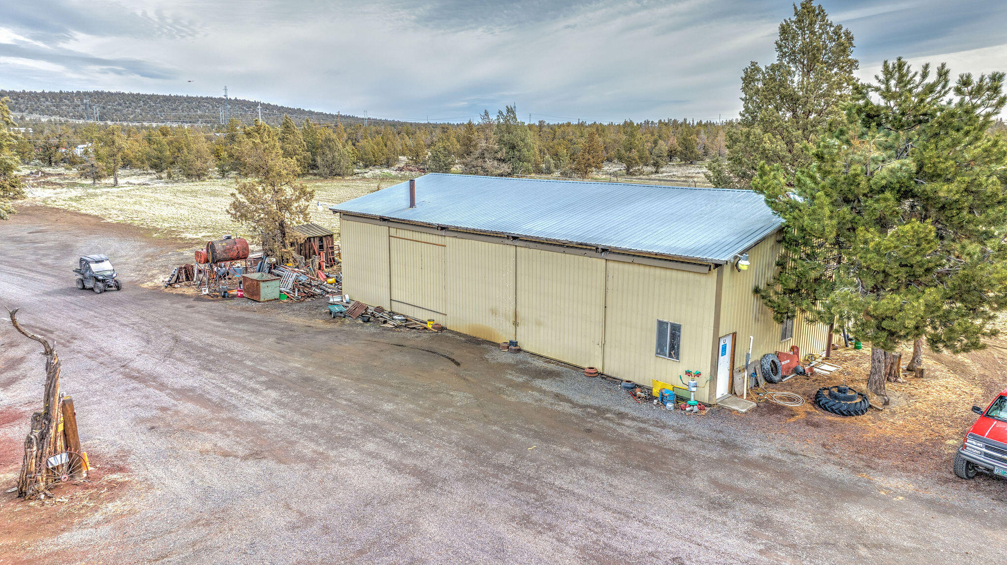 Large tan ranch workshop with a metal roof in a gravel yard among evergreens and scattered equipment.