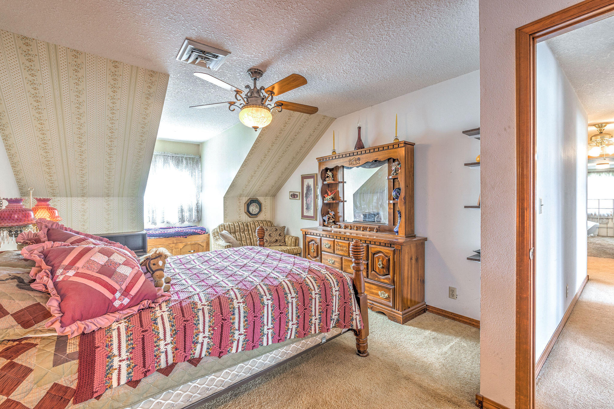Furnished bedroom with colorful patchwork quilts, sloped patterned walls, and an ornate wooden dresser with mirror.