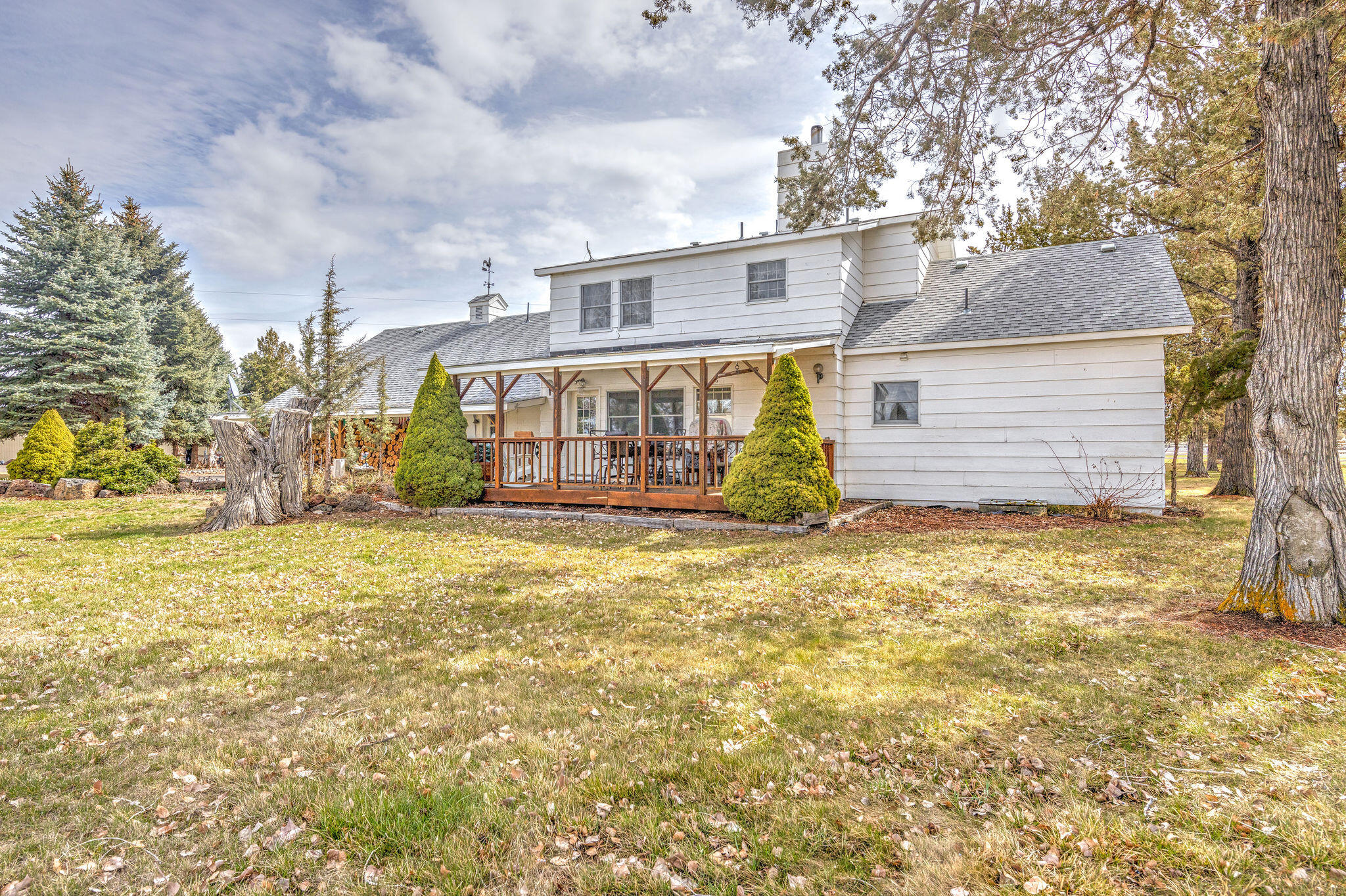 White two-story house with a covered wooden porch, framed by mature trees and a leaf-strewn lawn.