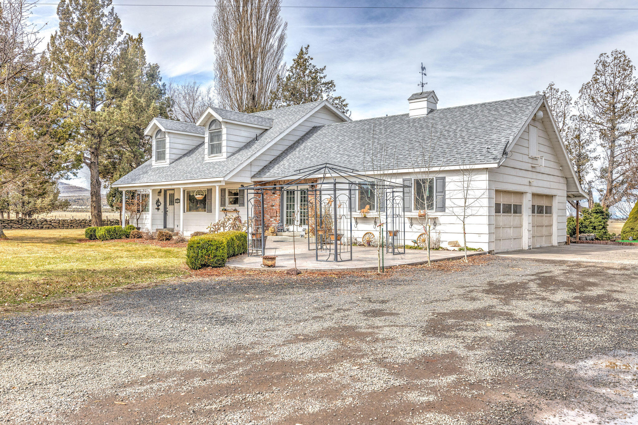 White farmhouse with dormer windows, attached garage, and metal garden arbor beside a gravel driveway.