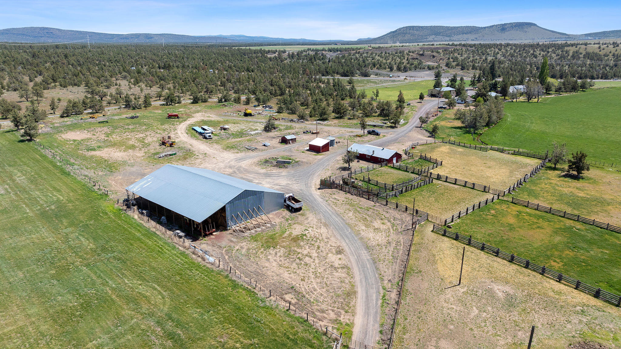 Aerial view of a ranch with a large barn, fenced paddocks, open pasture, and distant hills.
