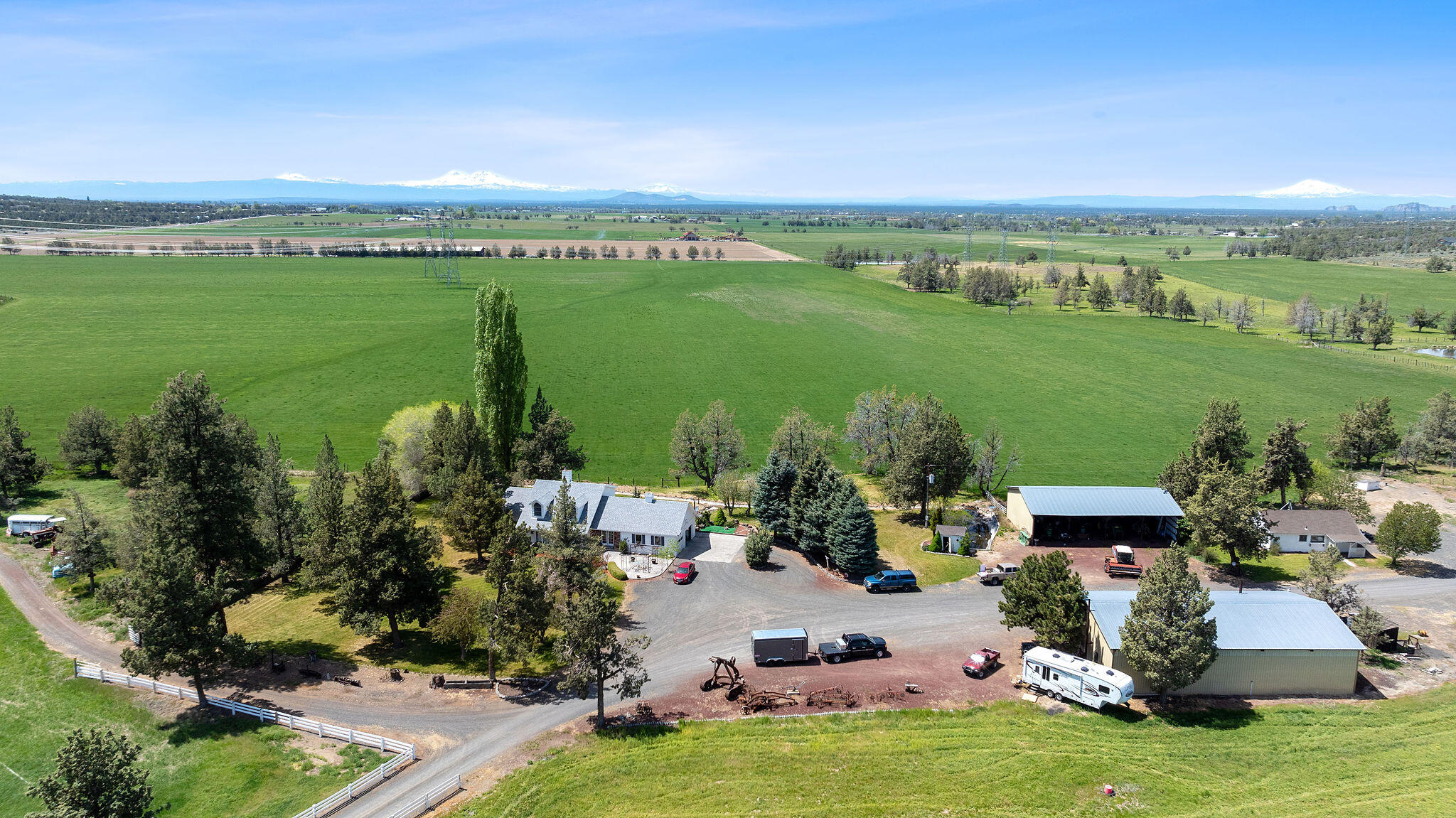Aerial view of a rural home and outbuildings amid green fields, with snow-capped mountains on the horizon.