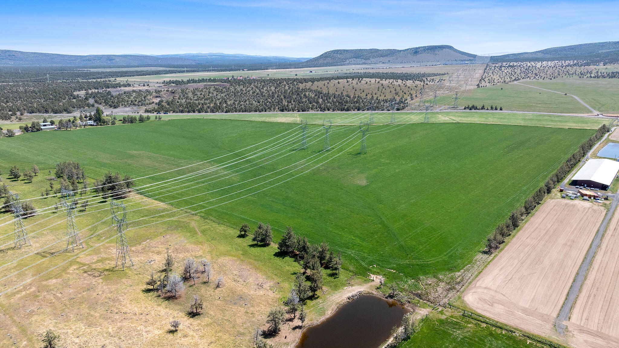 Aerial farmland crossed by power lines, with a pond and distant hills.