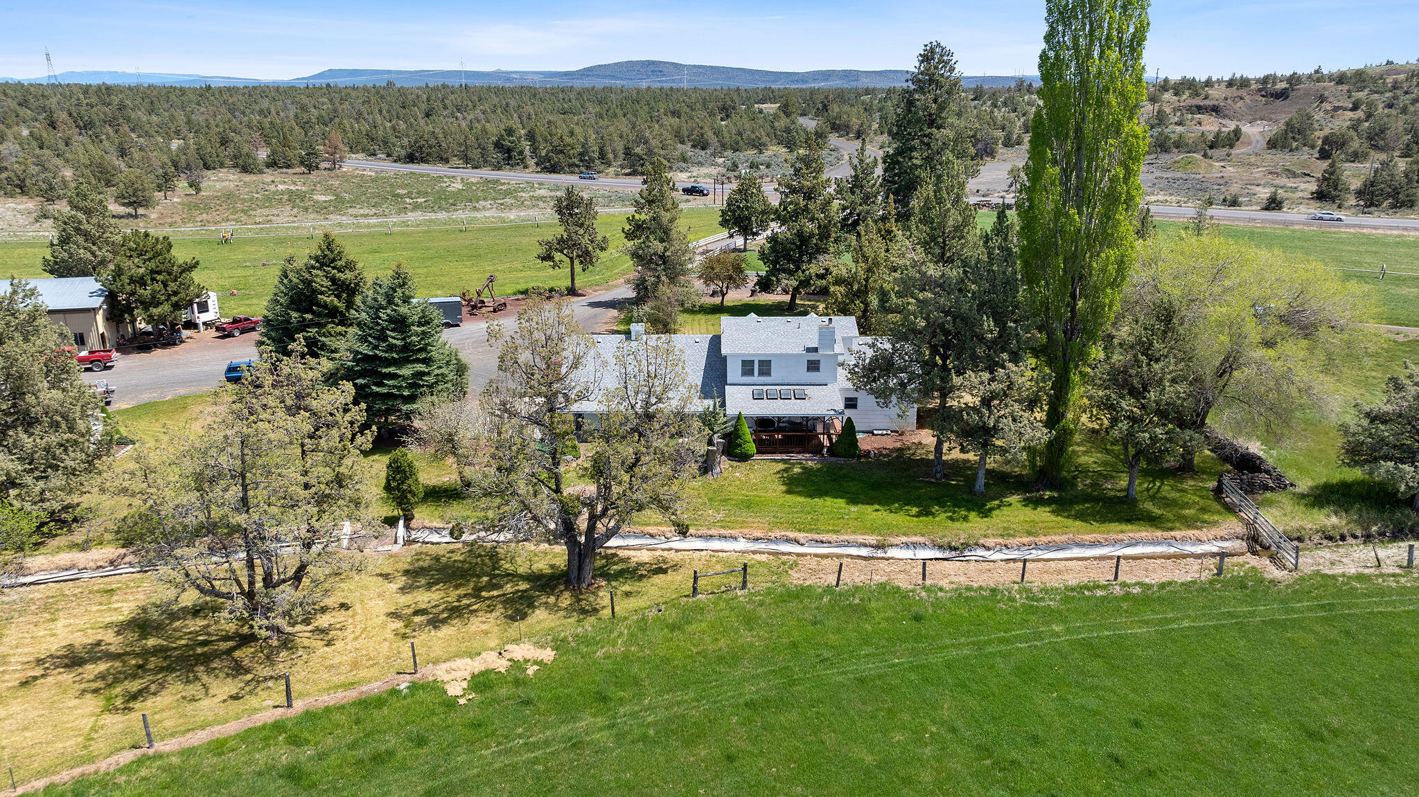 Aerial view of a white two-story home surrounded by mature trees, green lawn, and rural outbuildings.