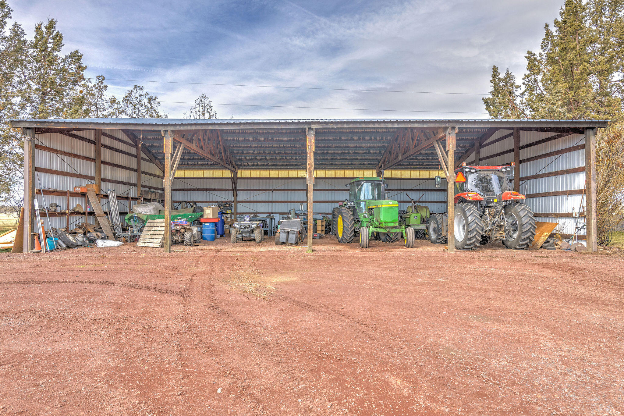 Open-sided farm equipment shed filled with tractors and machinery on a gravel lot.