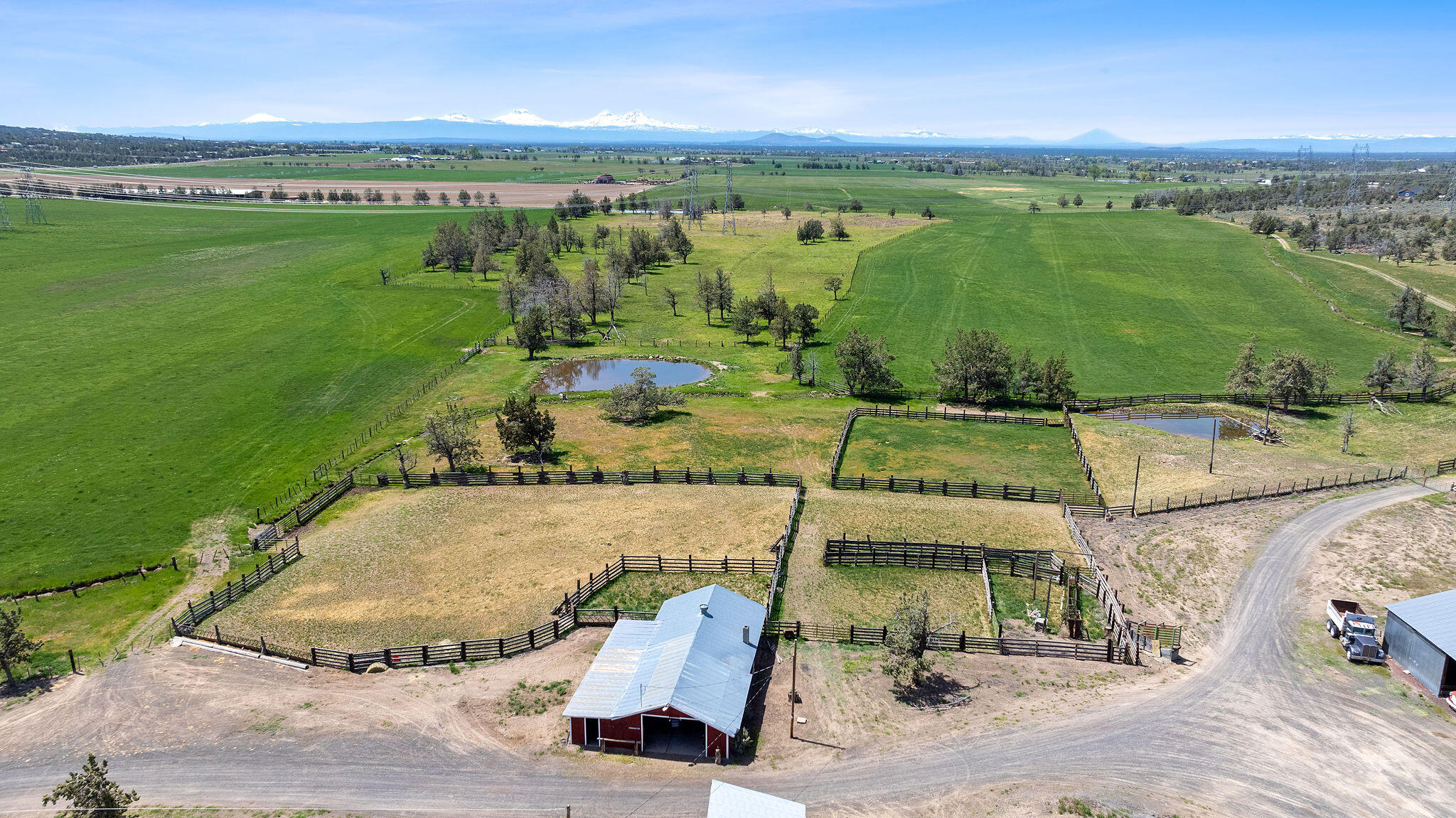 Aerial view of fenced ranch pastures and ponds, with a red barn and snow-capped mountains beyond.