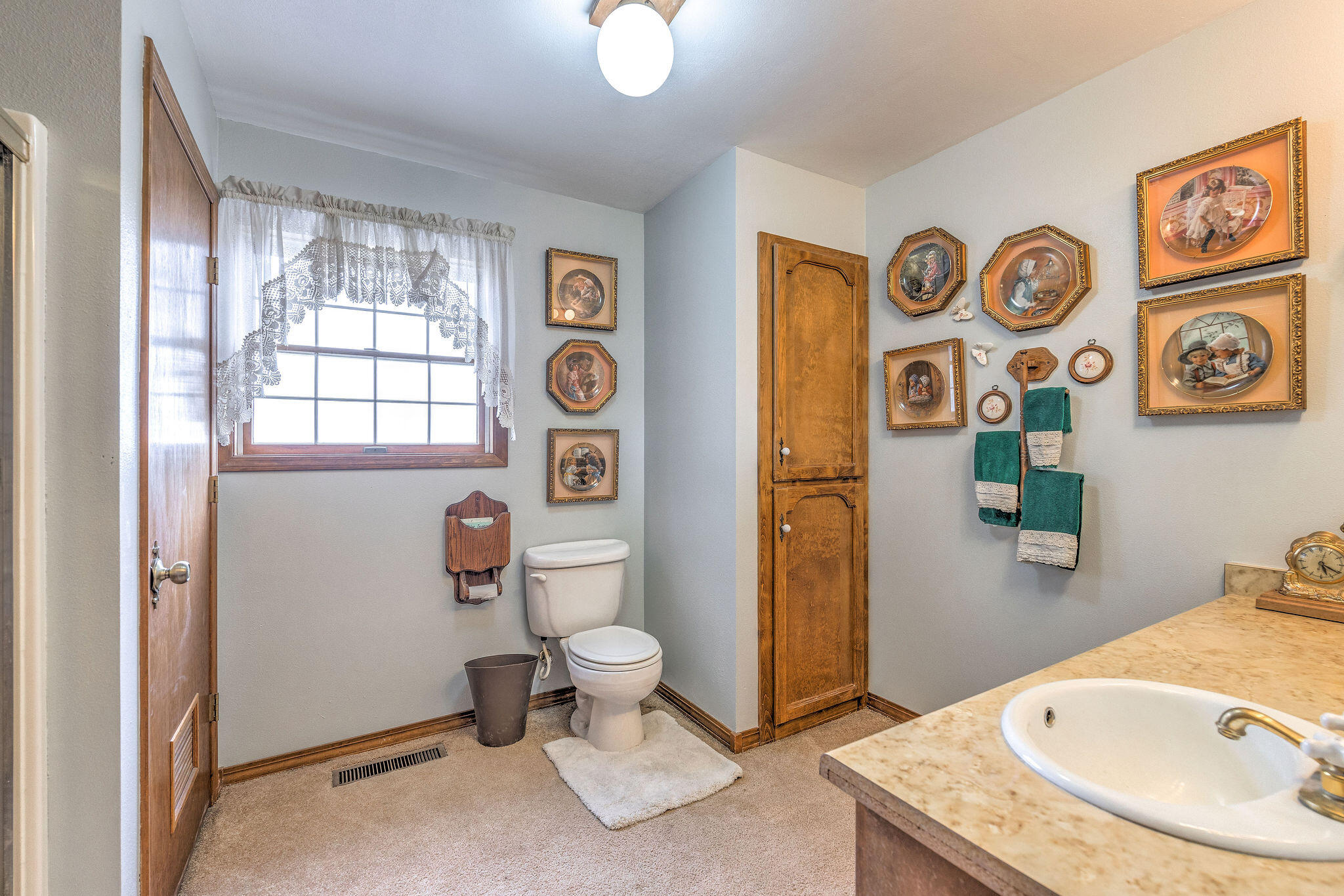 Bathroom with a lace-curtained window, toilet, wood cabinetry, and beige sink countertop.