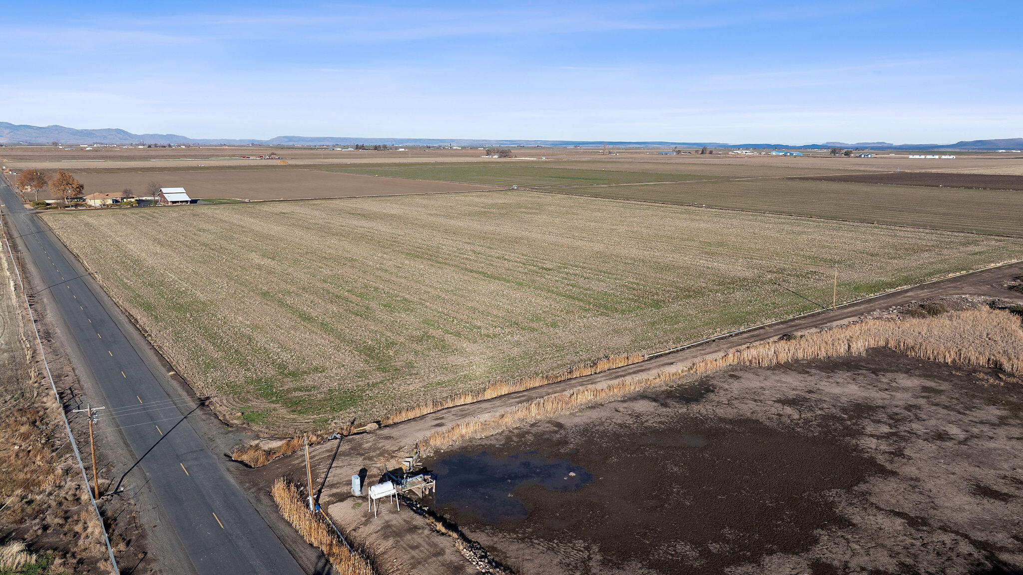 Aerial view of vacant farmland beside a paved road, with farm buildings and distant hills.