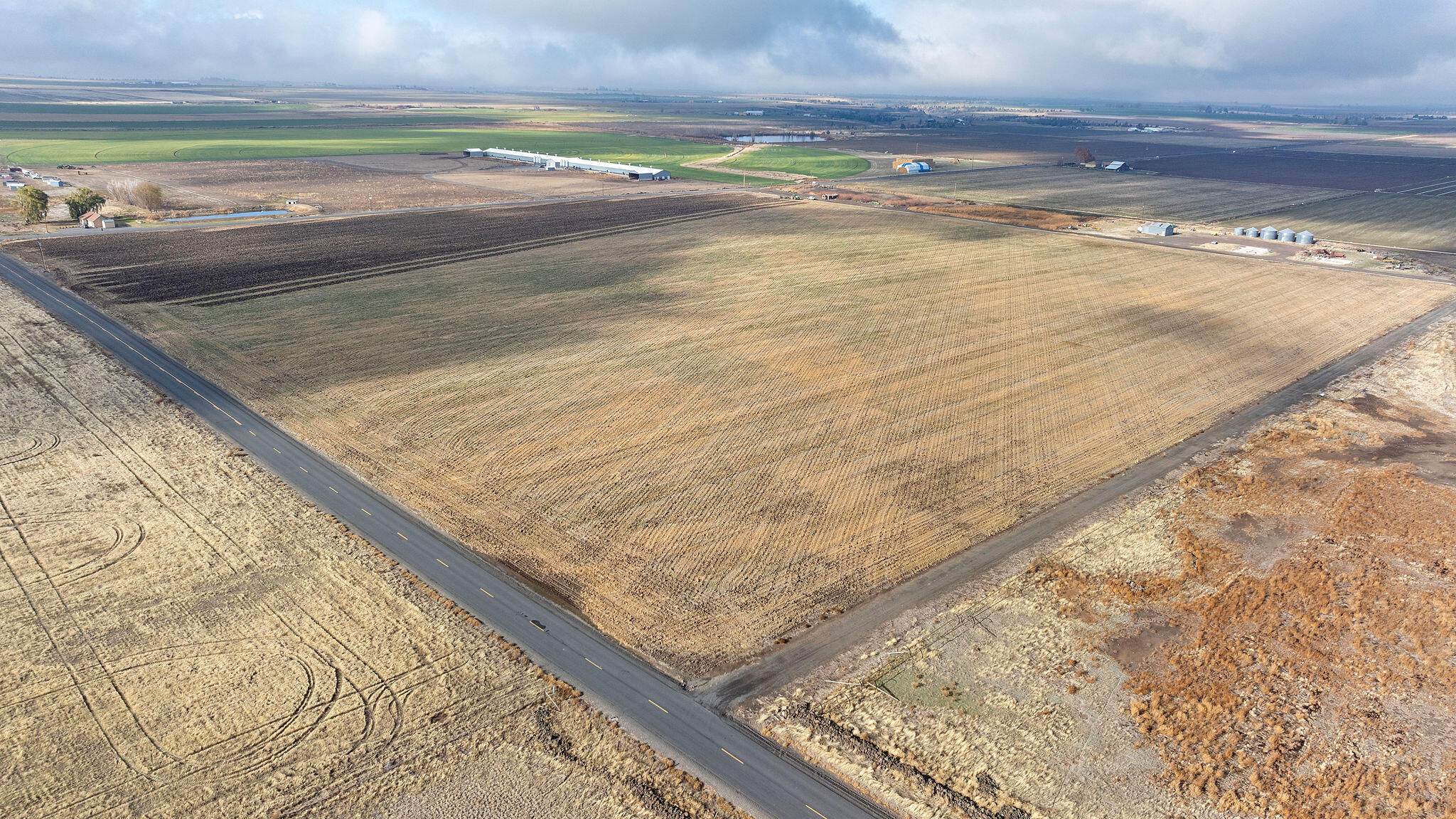 Aerial view of expansive farm fields divided by roads, with farm buildings and green plots in the distance.