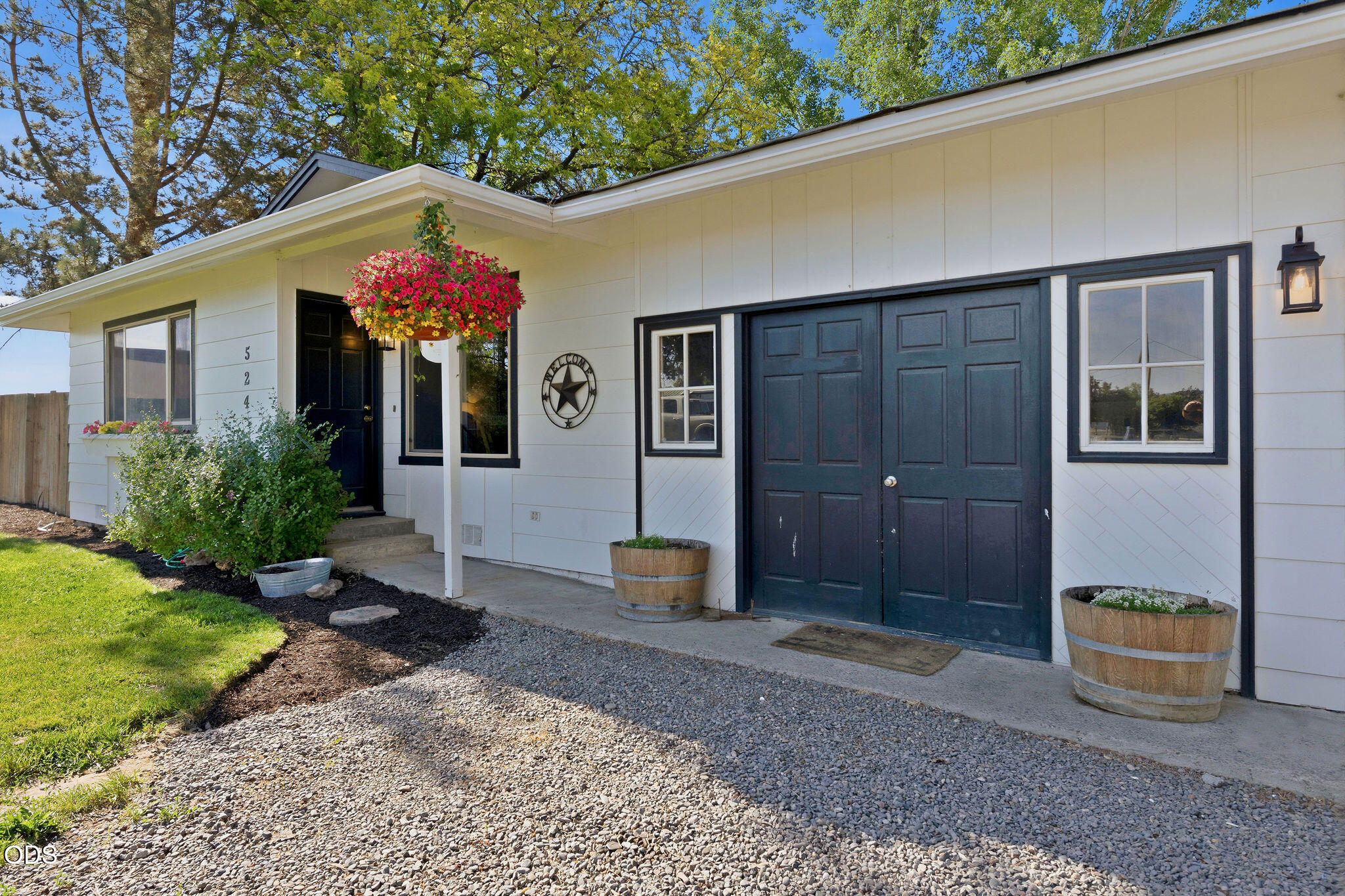 White house with dark blue garage doors, covered entry, colorful hanging flowers, and gravel driveway.