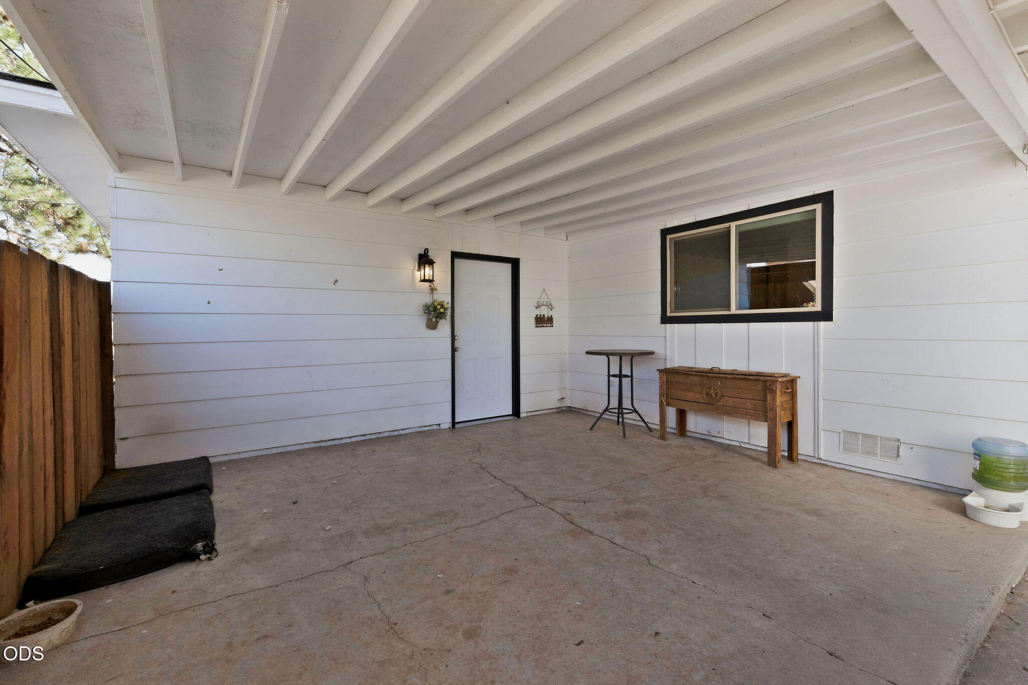 Covered porch with white siding, exposed ceiling beams, and a wooden chest beside the window.