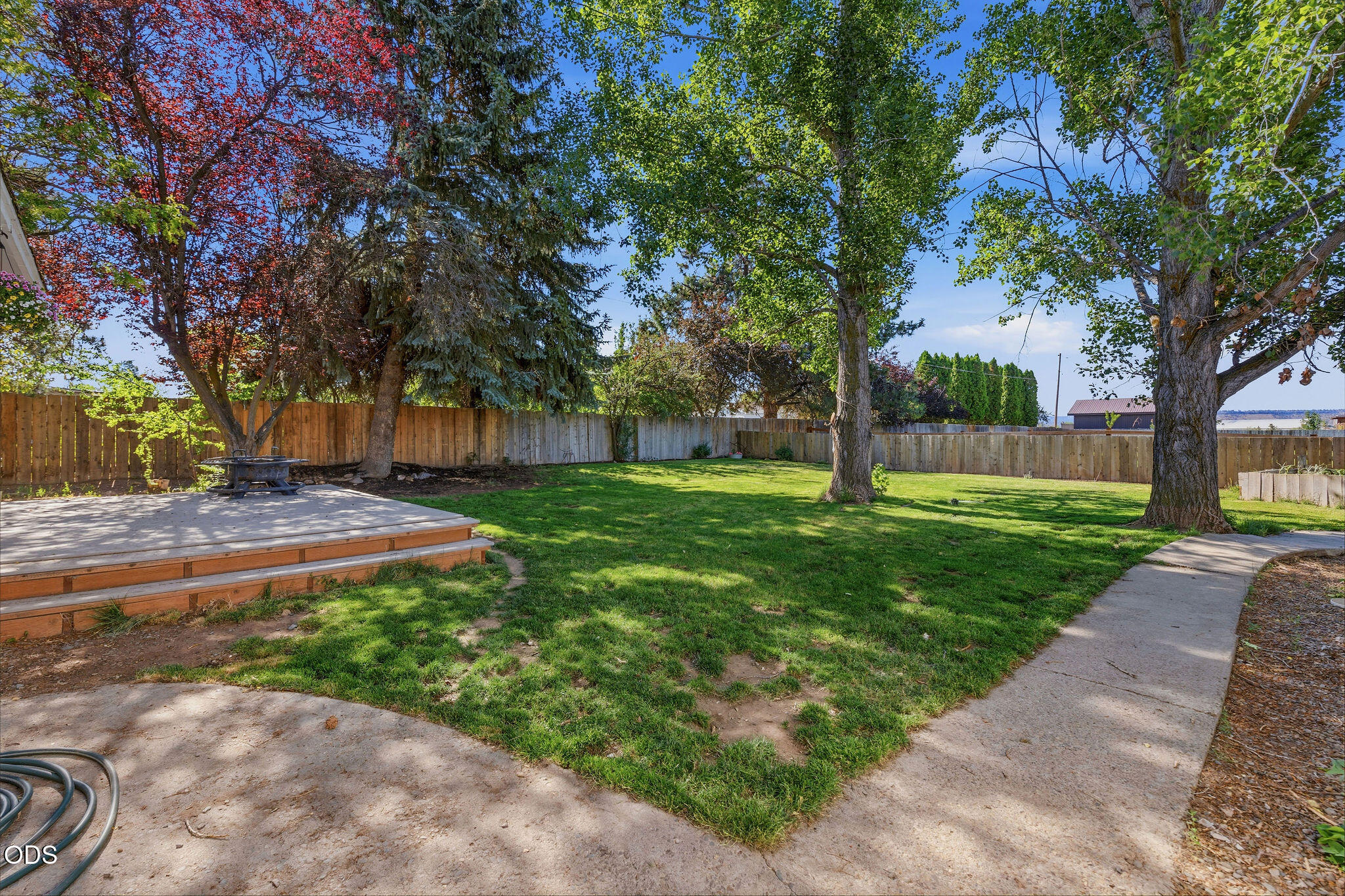 Fenced backyard with a green lawn, mature trees, raised deck, fire pit, and concrete paths.