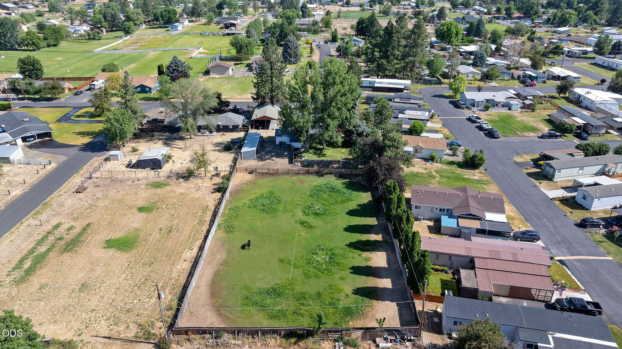 Aerial view of a fenced property with a broad grassy yard, open ground, and neighboring homes.