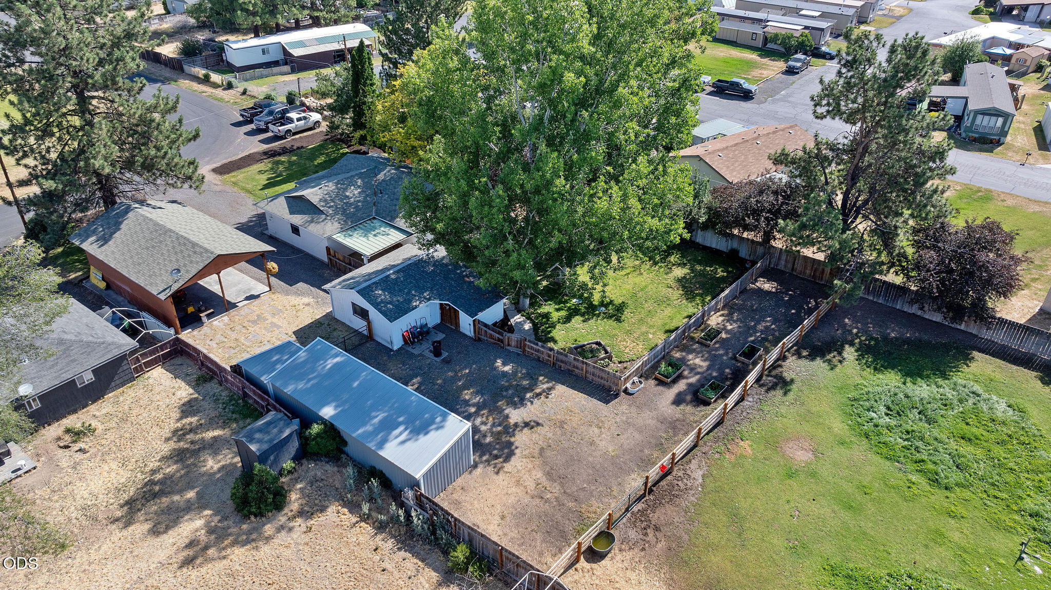 Aerial view of the property’s fenced yard, outbuildings, garden beds, and mature trees.