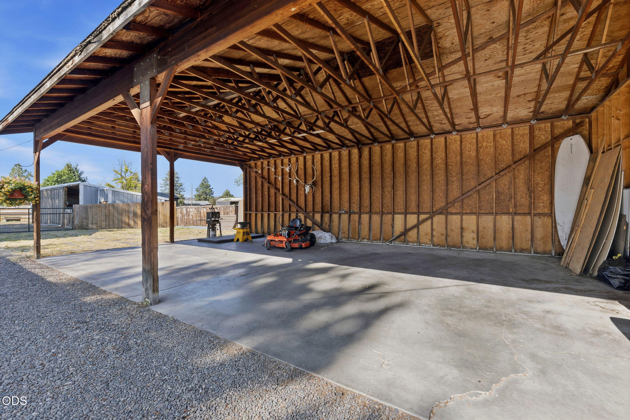 Open-sided wooden barn with exposed roof trusses, concrete floor, and a riding mower.