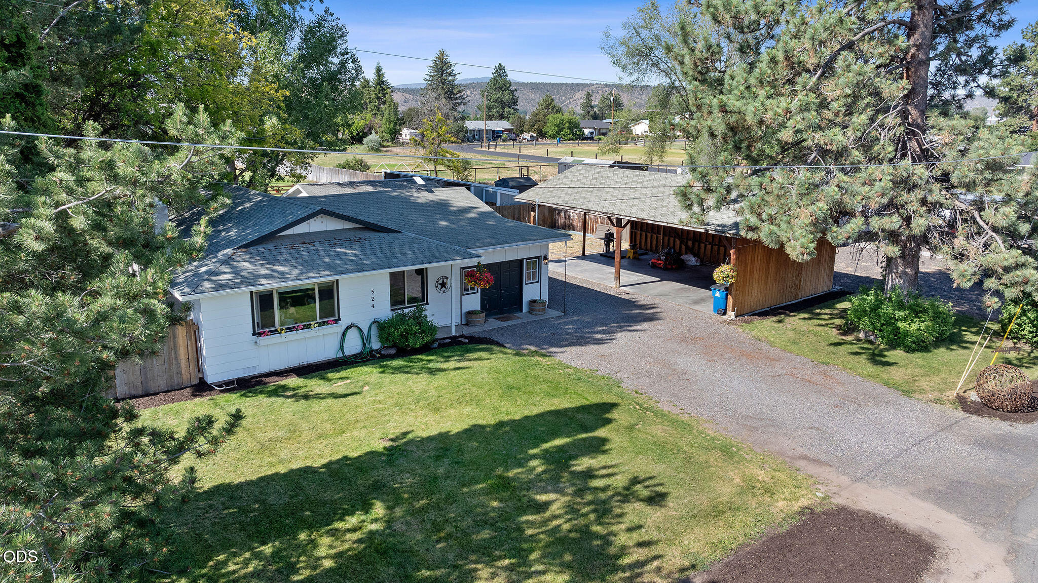 Single-story white home and covered carport amid pine trees, lawn, and distant mountain views.