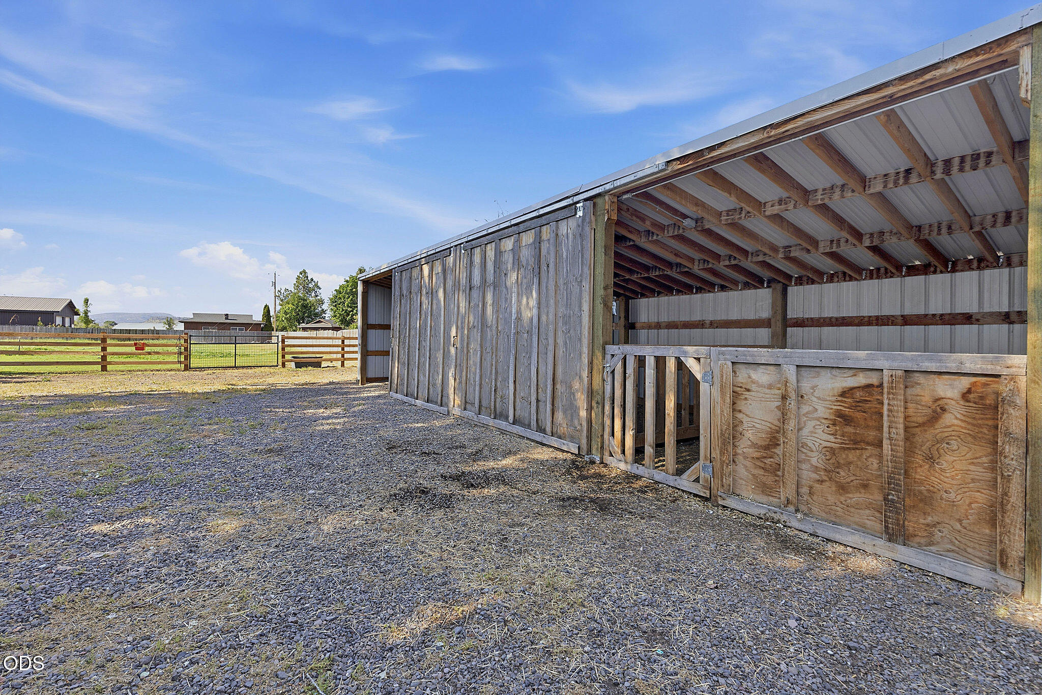 Open-sided wooden livestock shelter beside a fenced gravel paddock.