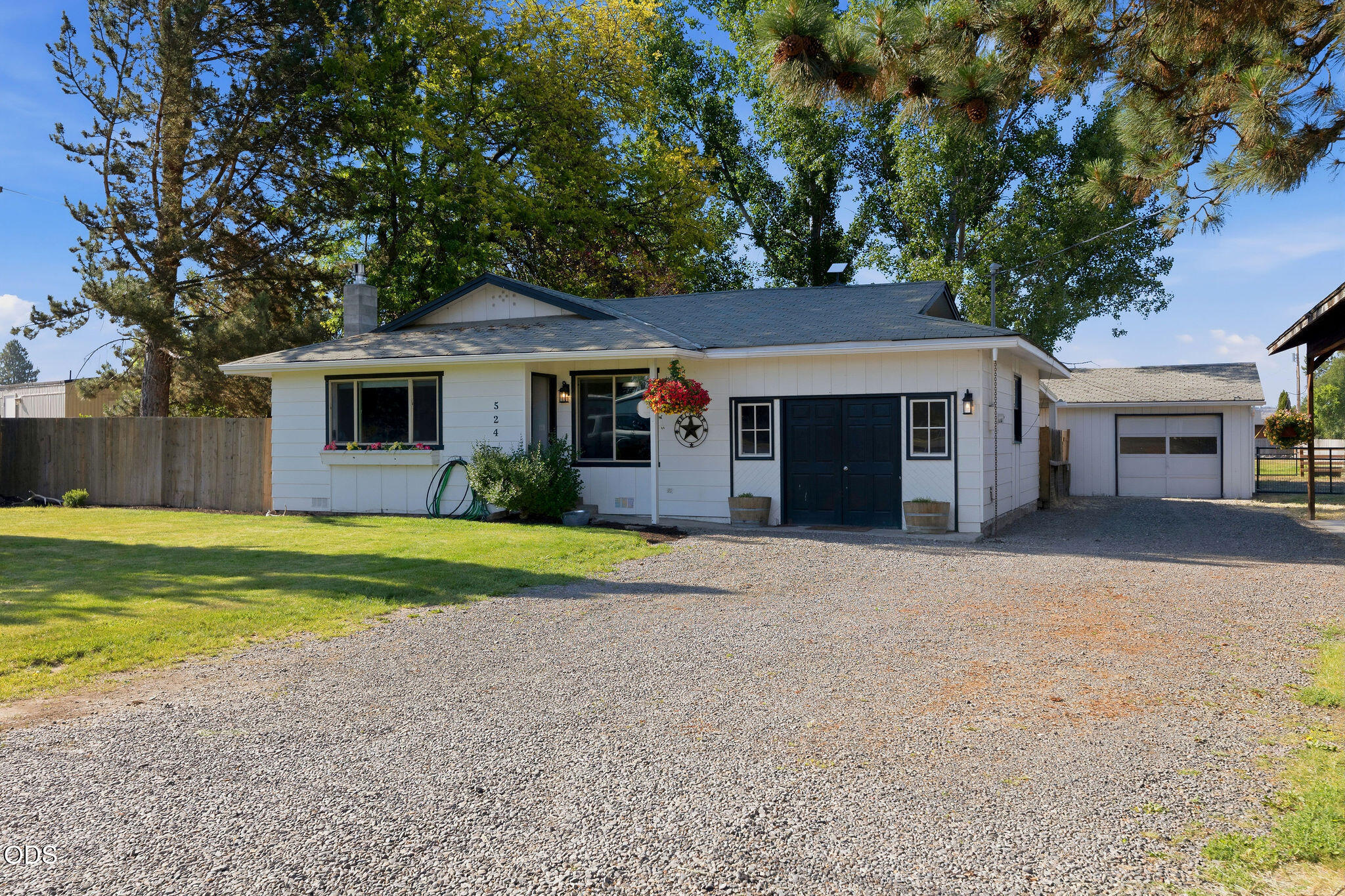 White single-story home with dark blue doors, gravel driveway, and detached garage beneath mature trees.