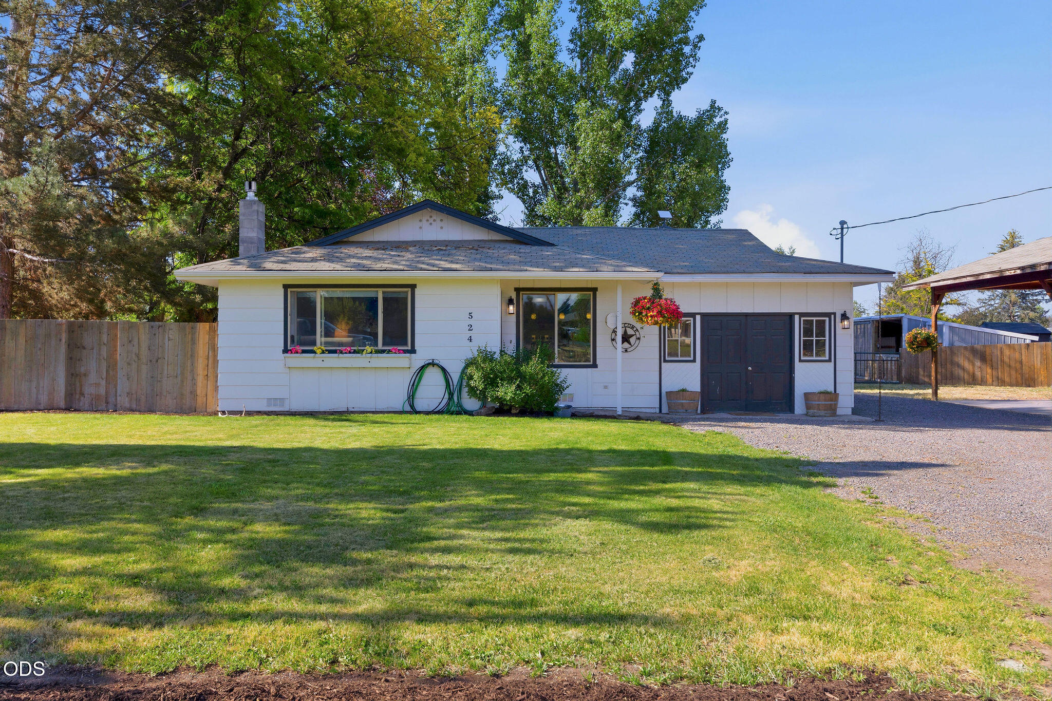 White single-story home with dark garage door, gravel driveway, and a shaded front lawn.