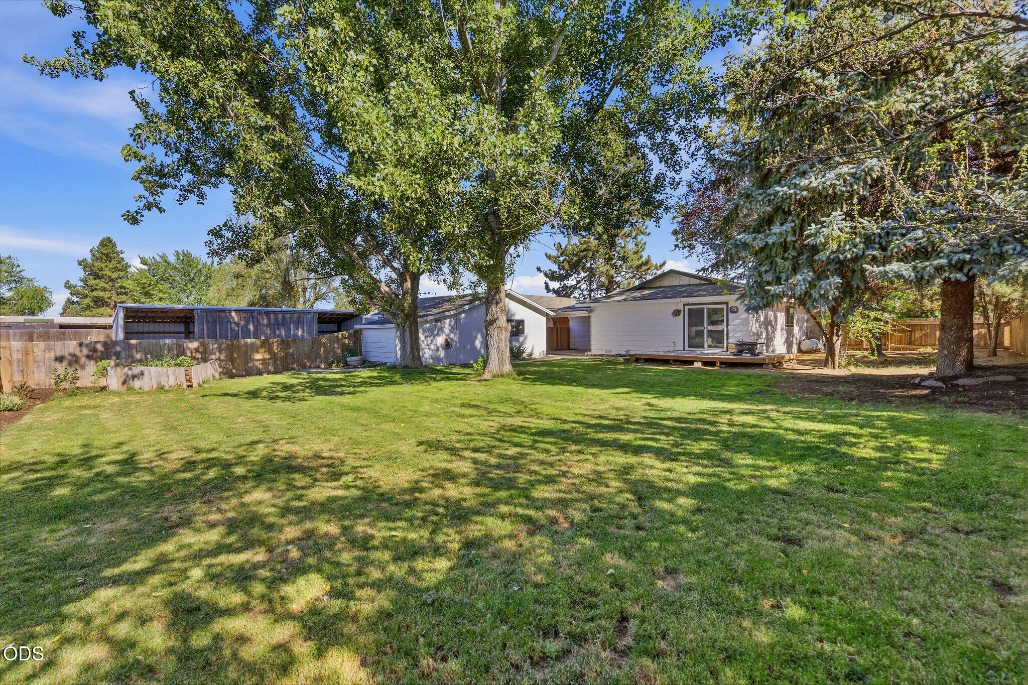 Single-story home behind a broad lawn, shaded by mature trees, with a wooden deck and fenced yard.