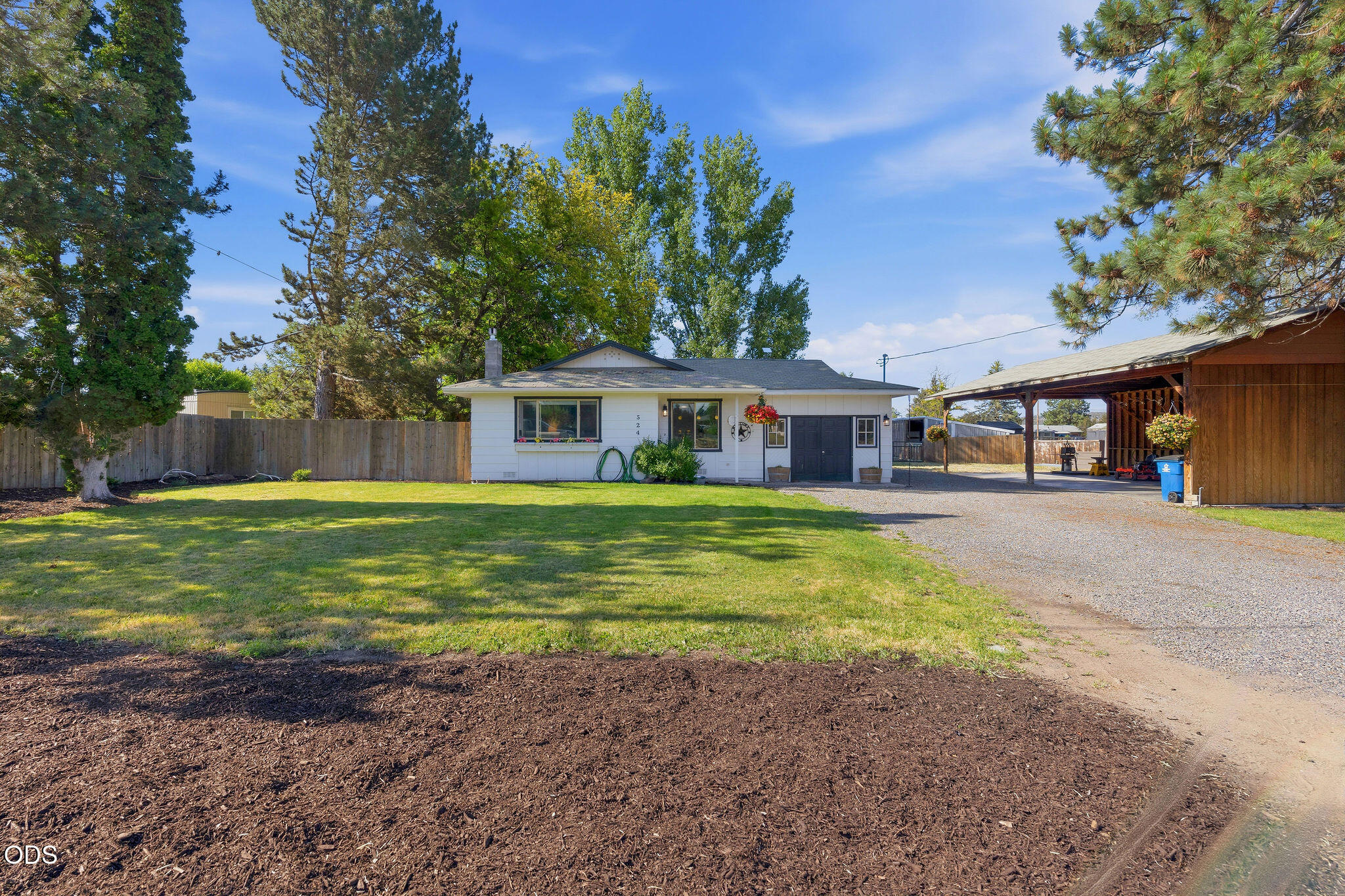 Single-story white house with a dark garage, gravel driveway, covered carport, and shaded lawn.