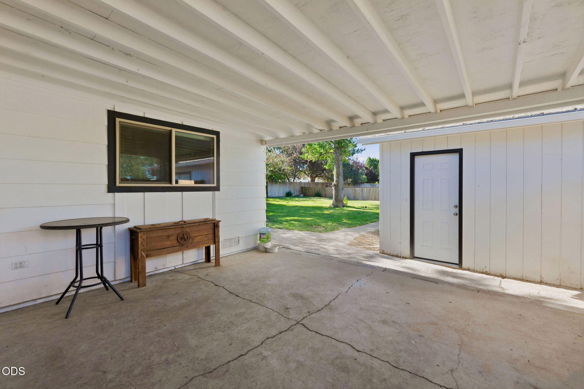 Covered concrete patio beside a white outbuilding, opening onto a fenced backyard with mature trees.