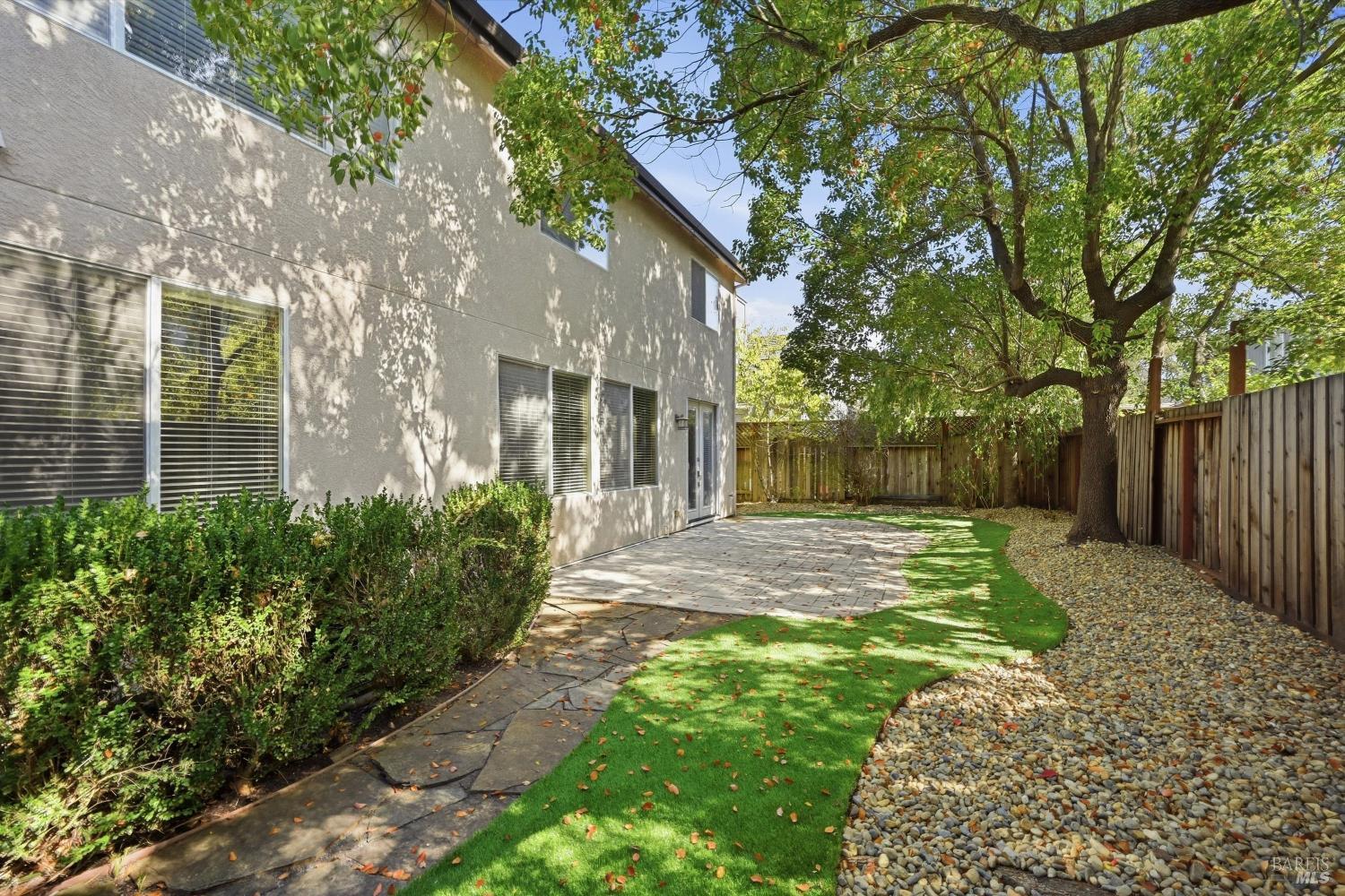House backyard with a paved patio, curved walkway, mature tree, and gravel landscaping.