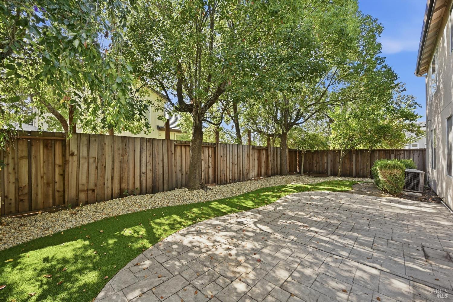 Backyard patio with stone pavers, green lawn, mature trees, and a wooden privacy fence.