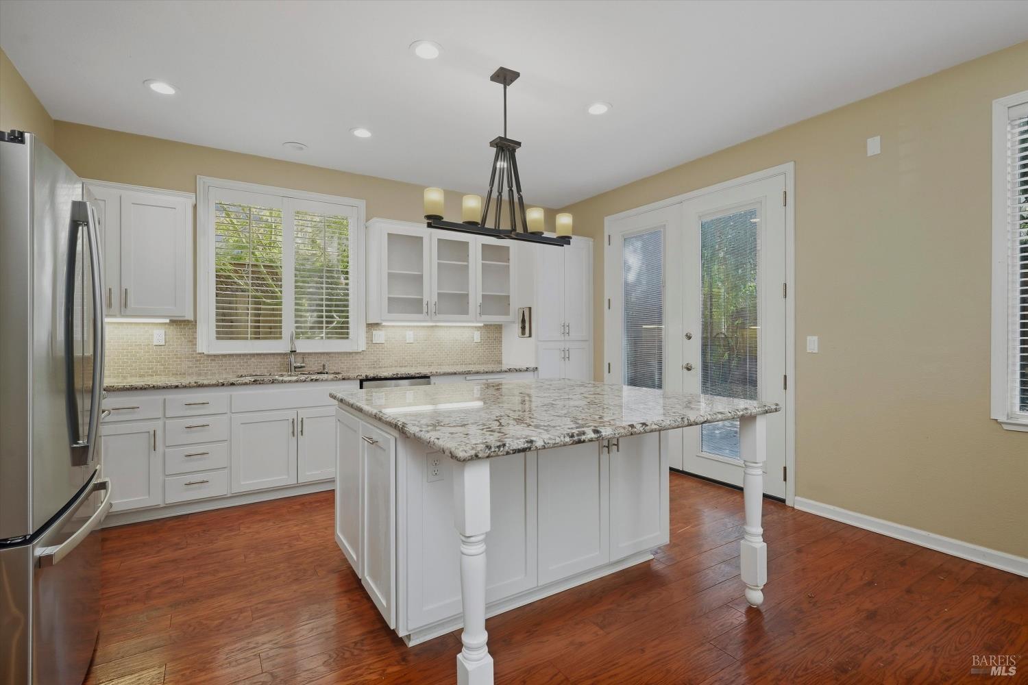 Kitchen with white cabinetry, a large granite island, stainless steel refrigerator, and reddish wood floors.