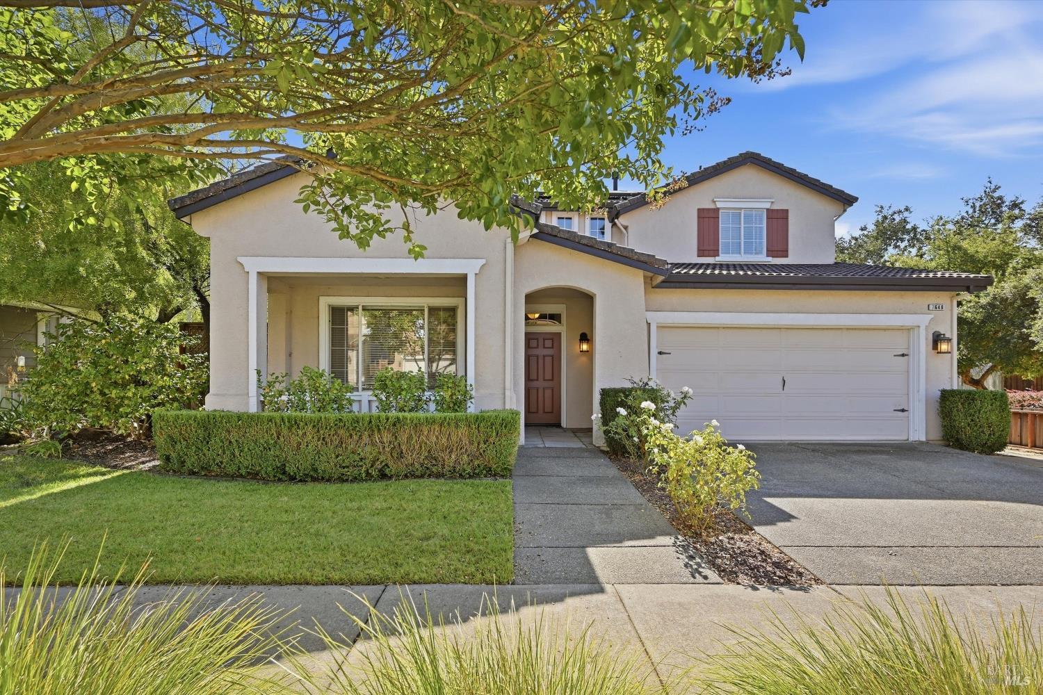 Two-story stucco home with a double garage, trimmed hedges, and leafy tree canopy.