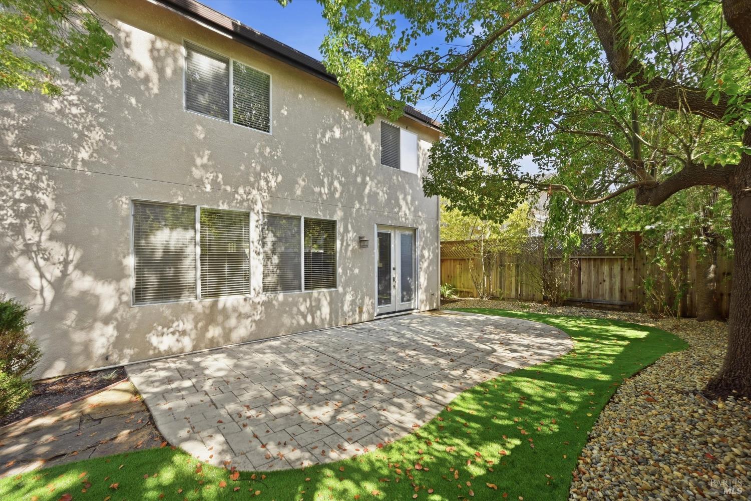 Home’s rear patio with pavers, sliding glass doors, lawn, and a large shade tree.