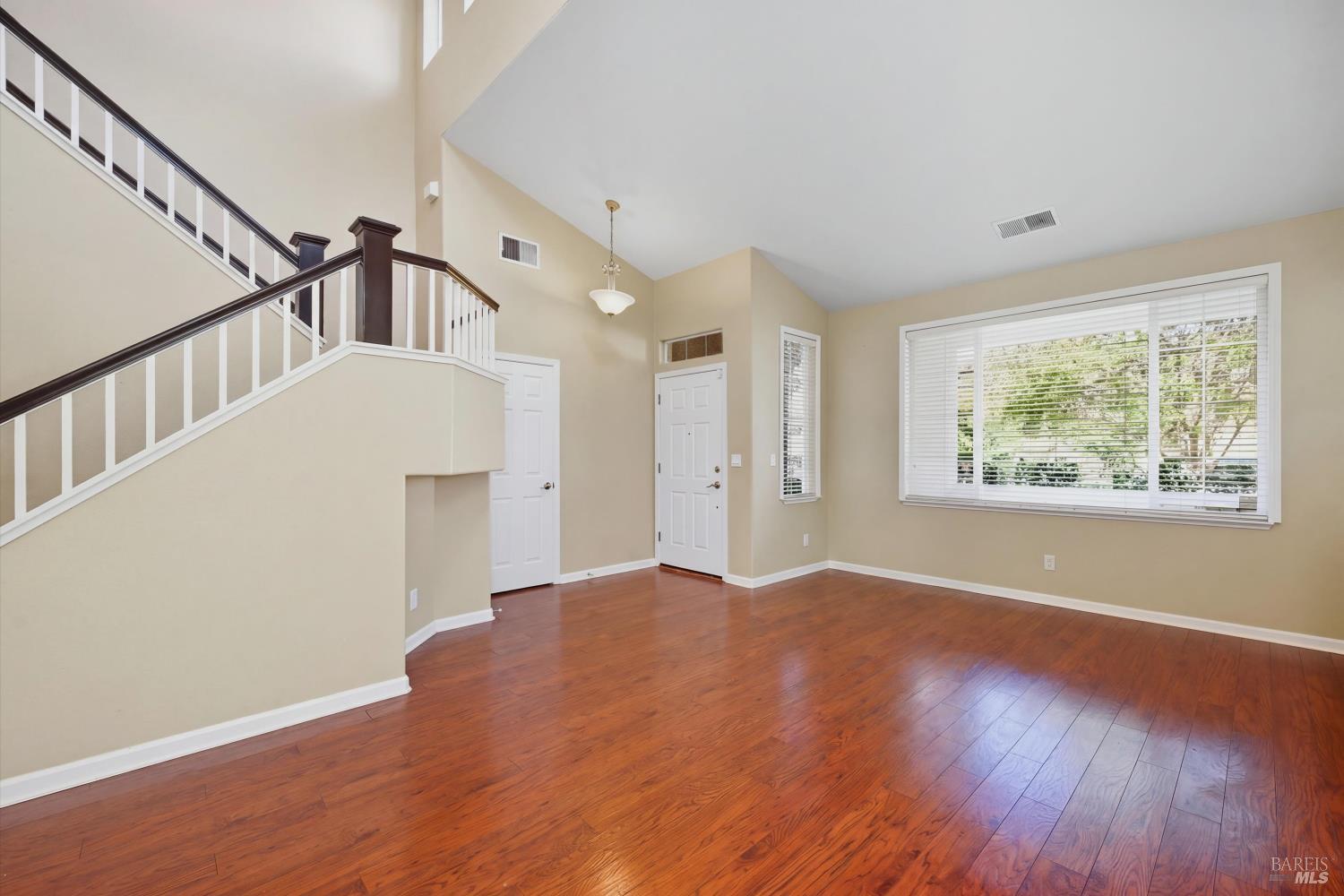 Empty living room with hardwood floors, a tall ceiling, and a staircase beside a large window.