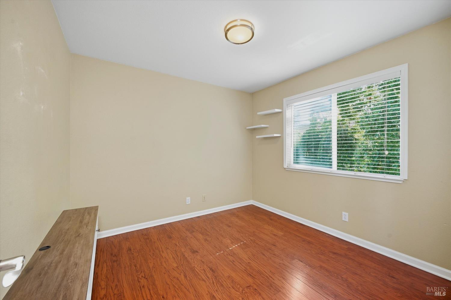 Empty bedroom with reddish hardwood floors, a large window with white blinds, and floating shelves.