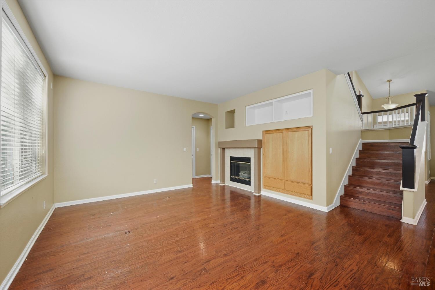 Empty living room with hardwood floors, a fireplace and built-in cabinet beside a dark wood staircase.