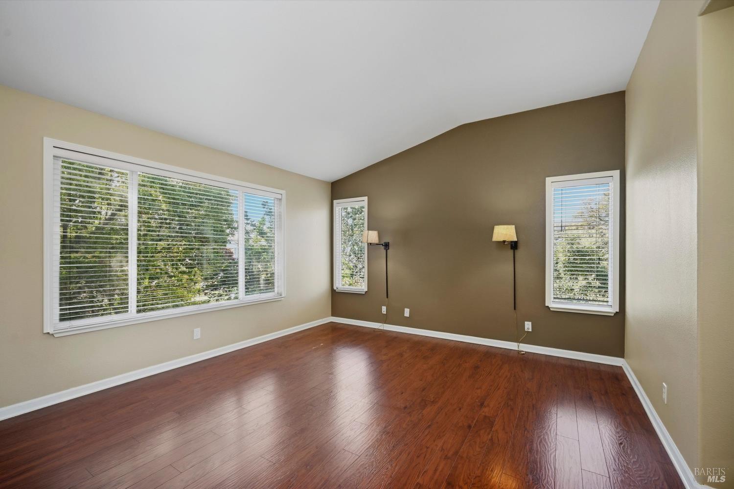 Empty room with reddish-brown wood flooring, a dark accent wall, and three windows with white blinds.