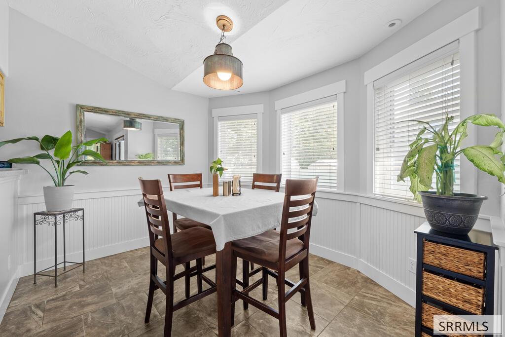 Dining nook with a dark wood table, three bright windows, white wainscoting, and potted plants.