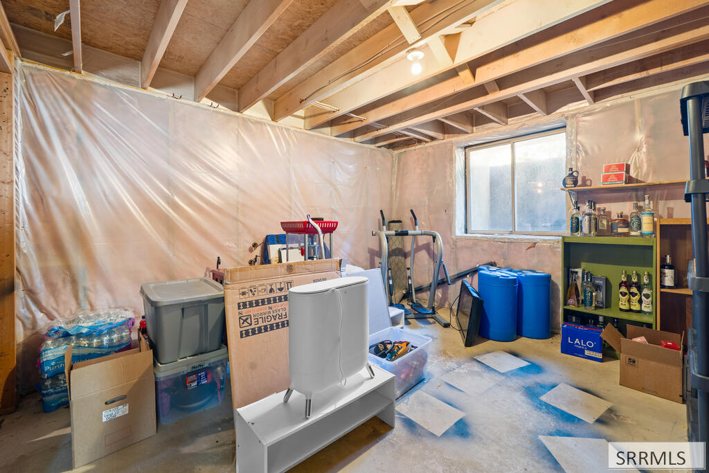 Unfinished basement with exposed joists, plastic-covered walls, stored belongings, exercise equipment, and a toilet on a cabinet.