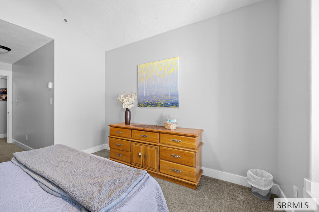 Bedroom with pale bedding, a wooden dresser, and colorful artwork on a gray wall.